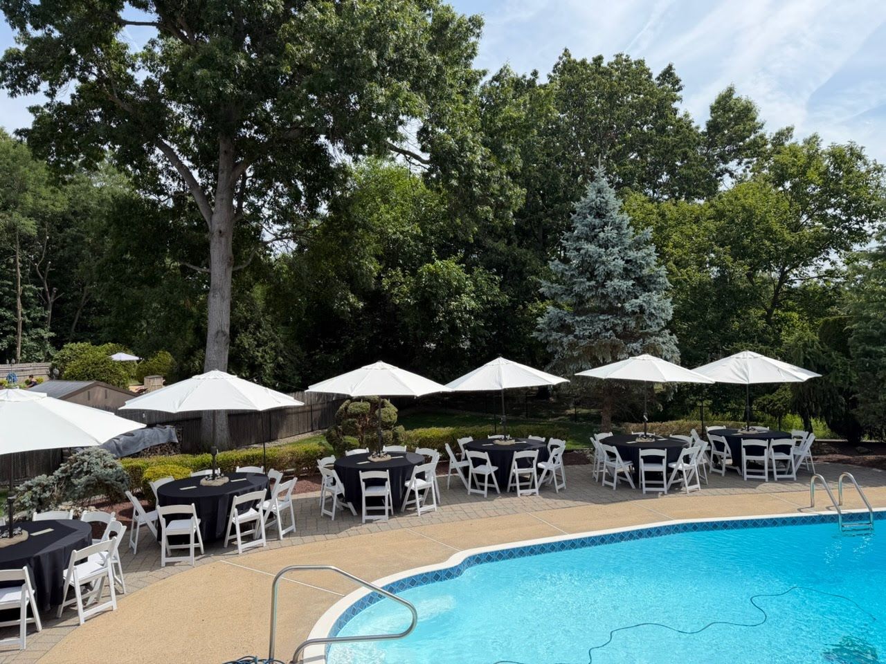 Six round tables in black linens with white padded chairs, each under a white market umbrella, along a backyard pool
