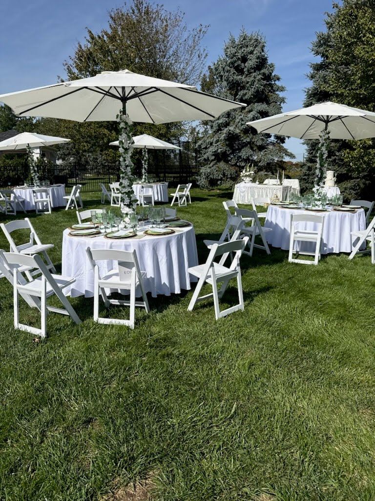 Round tables in white linens with white padded chairs under white market umbrellas, poles wrapped in white flowers and greenery